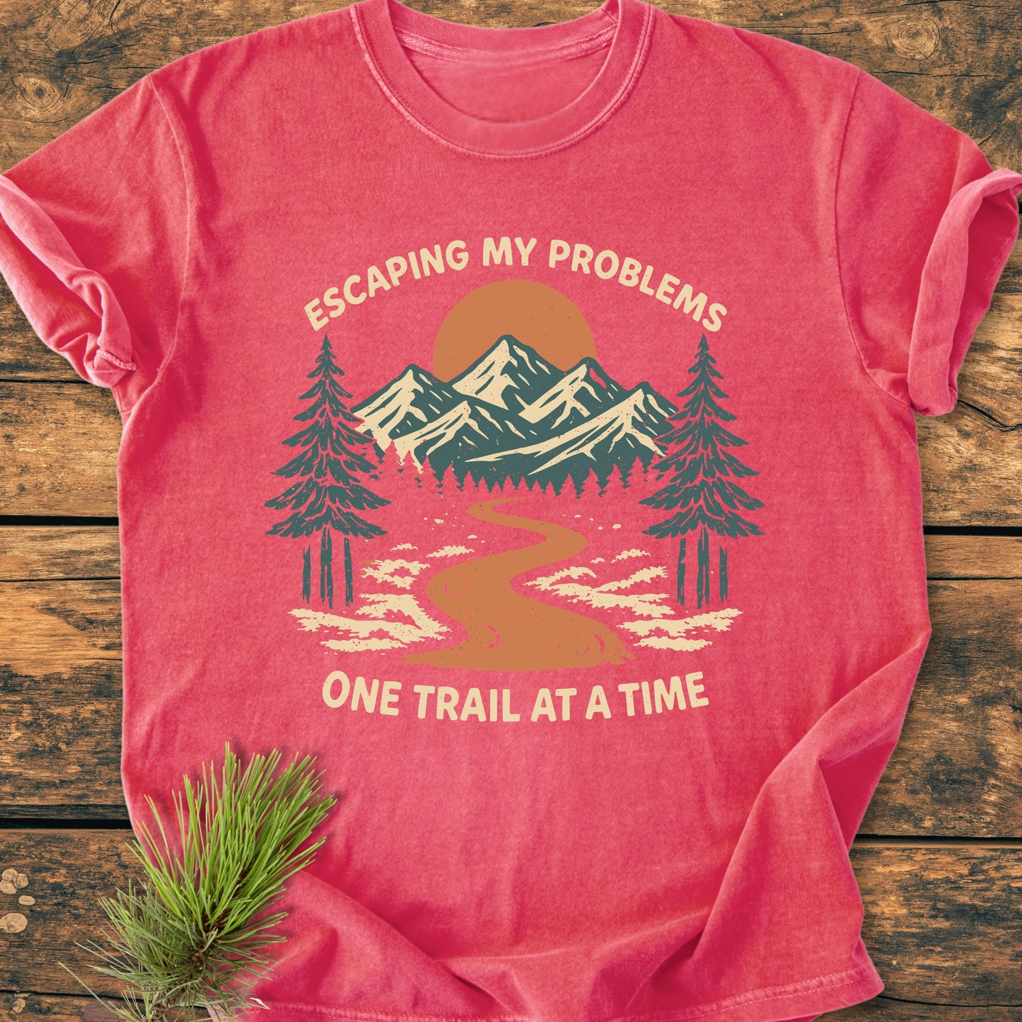 Escaping My Problems - Vintage Wash Tee: Red-coral T-shirt with an outdoors scene of trees and mountains, featuring the slogan. Laid flat on wood, accompanied by a small pine sprig.