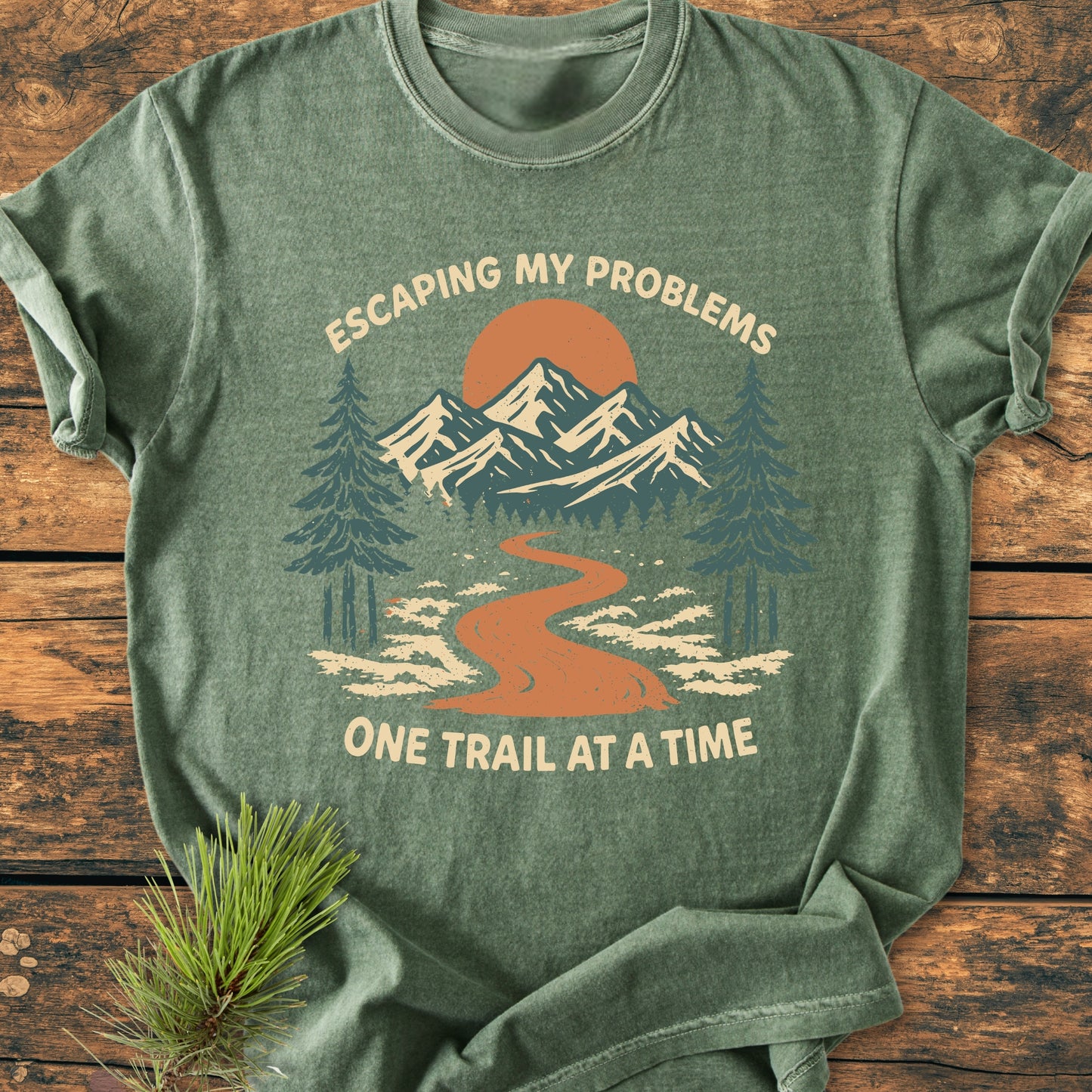 Escaping My Problems - Vintage Wash Tee, featuring a stylized outdoor scene with a trail, trees, and mountains, is displayed on a wooden surface, highlighting its relaxed, durable design and soft, garment-dyed fabric.