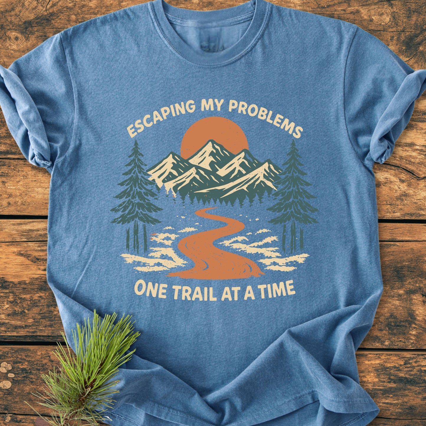 Escaping My Problems - Vintage Wash Tee: Heather-blue shirt with a scenic outdoors print, featuring mountains, trail, and sun, accompanied by motivational text. Laid on wood, accented with pine needles.