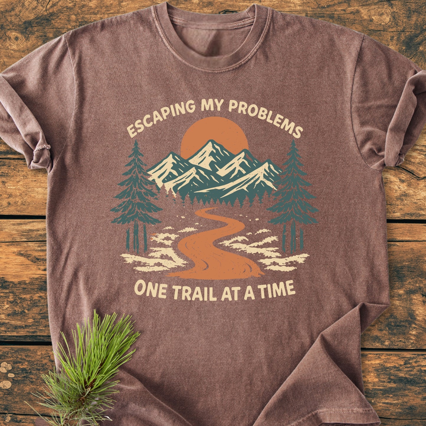 Escaping My Problems - Vintage Wash Tee features a mountain trail graphic with pine trees and sun, laid flat on wood, emphasizing outdoor adventure. Includes pine sprig for a nature-inspired touch.