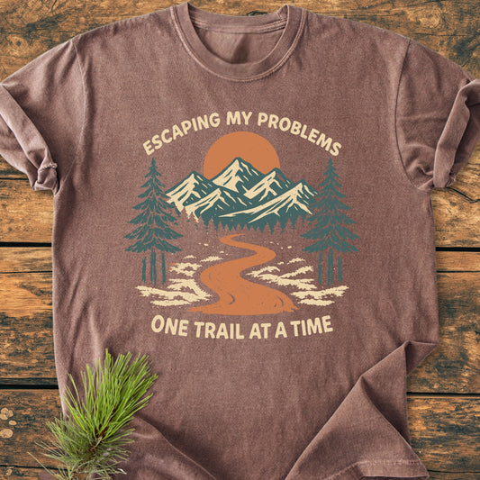 Escaping My Problems - Vintage Wash Tee features a mountain trail graphic with pine trees and sun, laid flat on wood, emphasizing outdoor adventure. Includes pine sprig for a nature-inspired touch.
