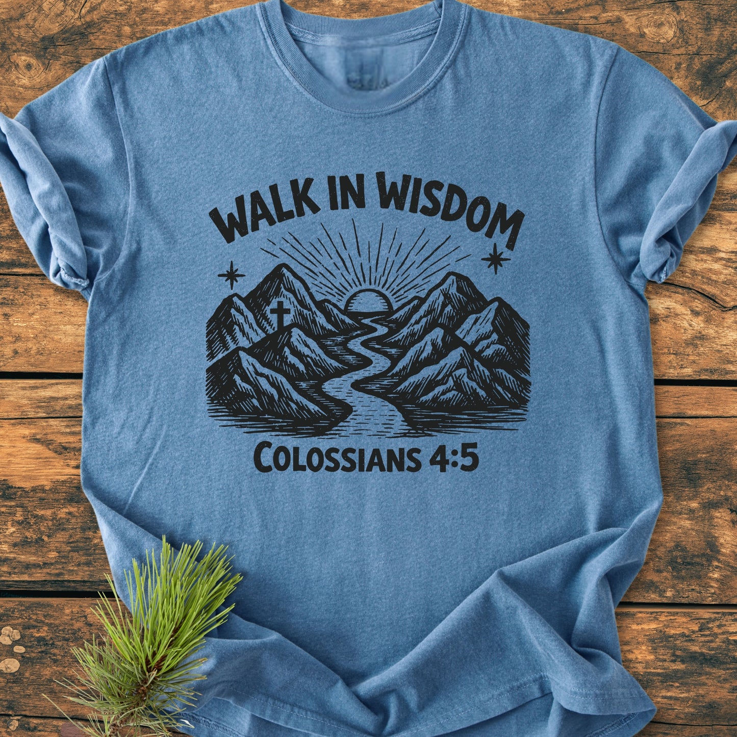 Walk In Wisdom, Colossians 4:5 - Vintage Wash Tee features a mountain graphic with a river and sun, laid flat on wood with a pine sprig, showcasing its relaxed fit and durable design.
