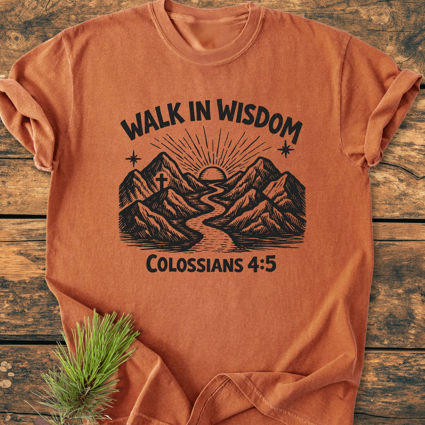 Walk In Wisdom, Colossians 4:5 - Vintage Wash Tee features a mountain landscape with a sun and stars, on a soft orange-rust color, laid flat with an evergreen sprig beside it.