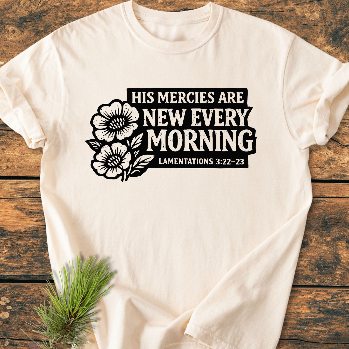 New Every Morning Lamentations 3:22-23 - Vintage Wash Tee displayed flat, features bold black text and floral illustrations on a light-cream shirt, styled with a pine sprig on a wooden surface.