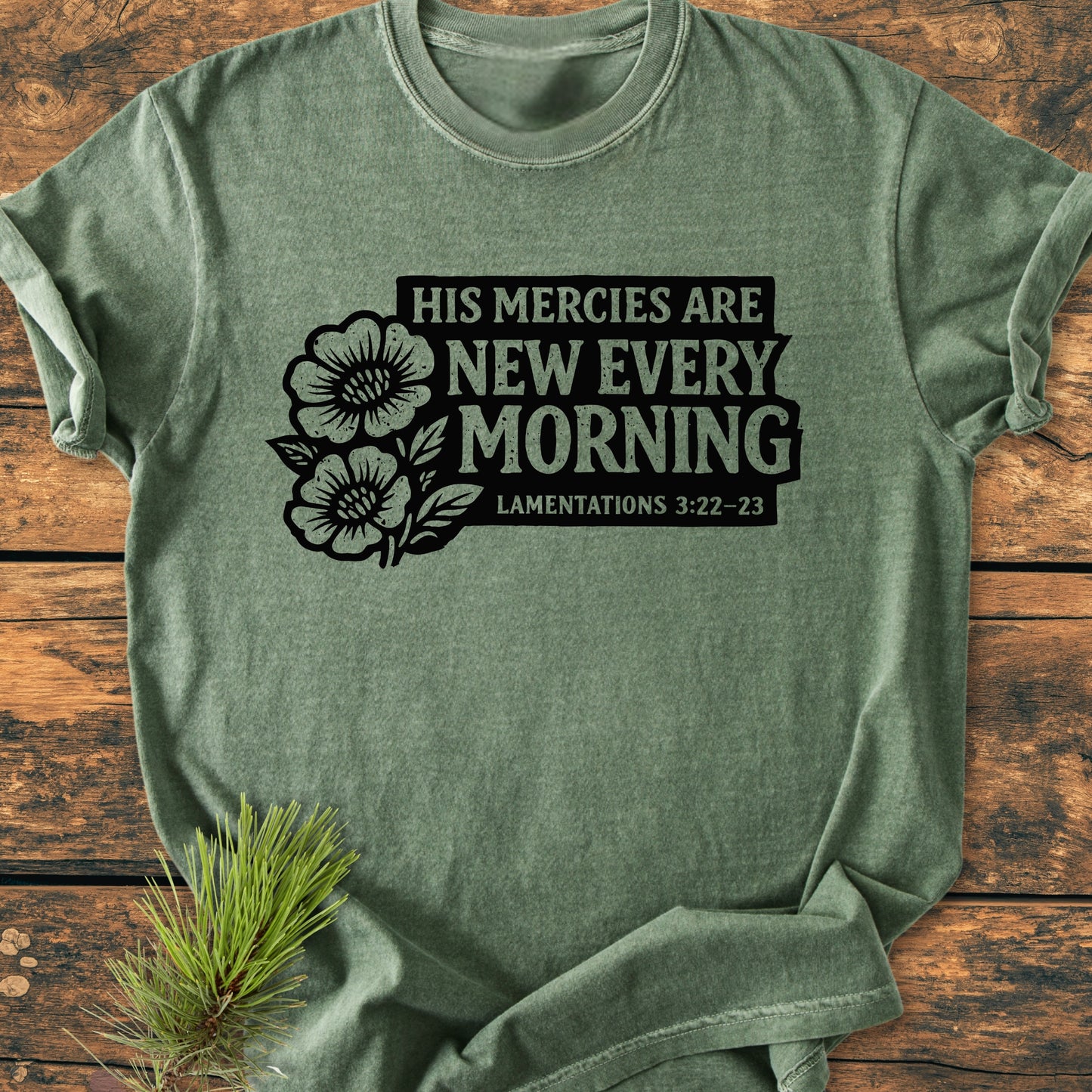 New Every Morning Lamentations 3:22-23 Vintage Wash Tee, heather-green with black floral scripture design, laid flat on wood, pine sprig accentuates earthy look, highlighting Comfort Colors' cozy, durable garment-dyed fabric.