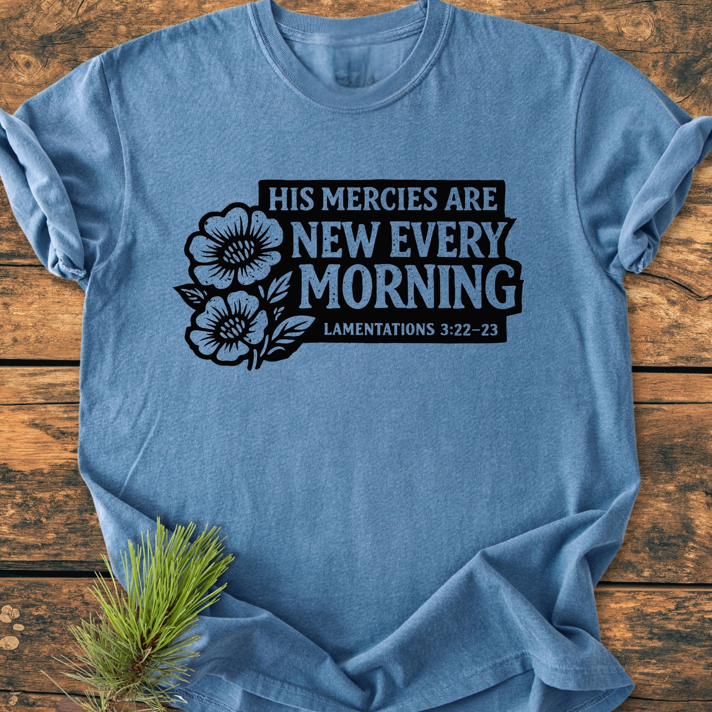 New Every Morning Lamentations 3:22-23 - Vintage Wash Tee features bold, black lettering of a Bible verse with flower graphics on a heather-blue T-shirt, styled with a pine sprig on a wooden surface.