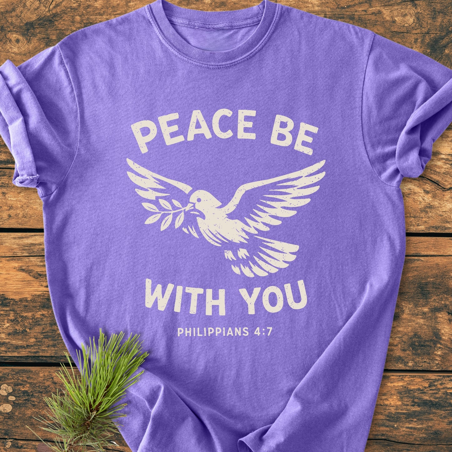 Peace Be With You Philippians 4:7 - Vintage Wash Tee features a heather-purple, soft-wrinkled T-shirt with a white dove graphic holding an olive branch, laid on a rustic wooden surface.