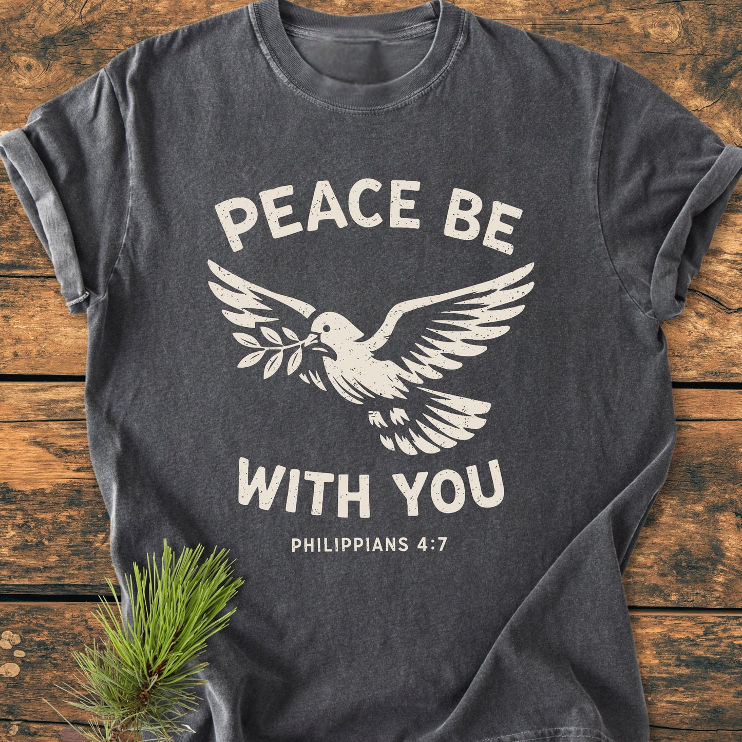 Peace Be With You Philippians 4:7 - Vintage Wash Tee features a stylized dove and message on charcoal-gray fabric, resting on a wooden surface with a decorative pine sprig.