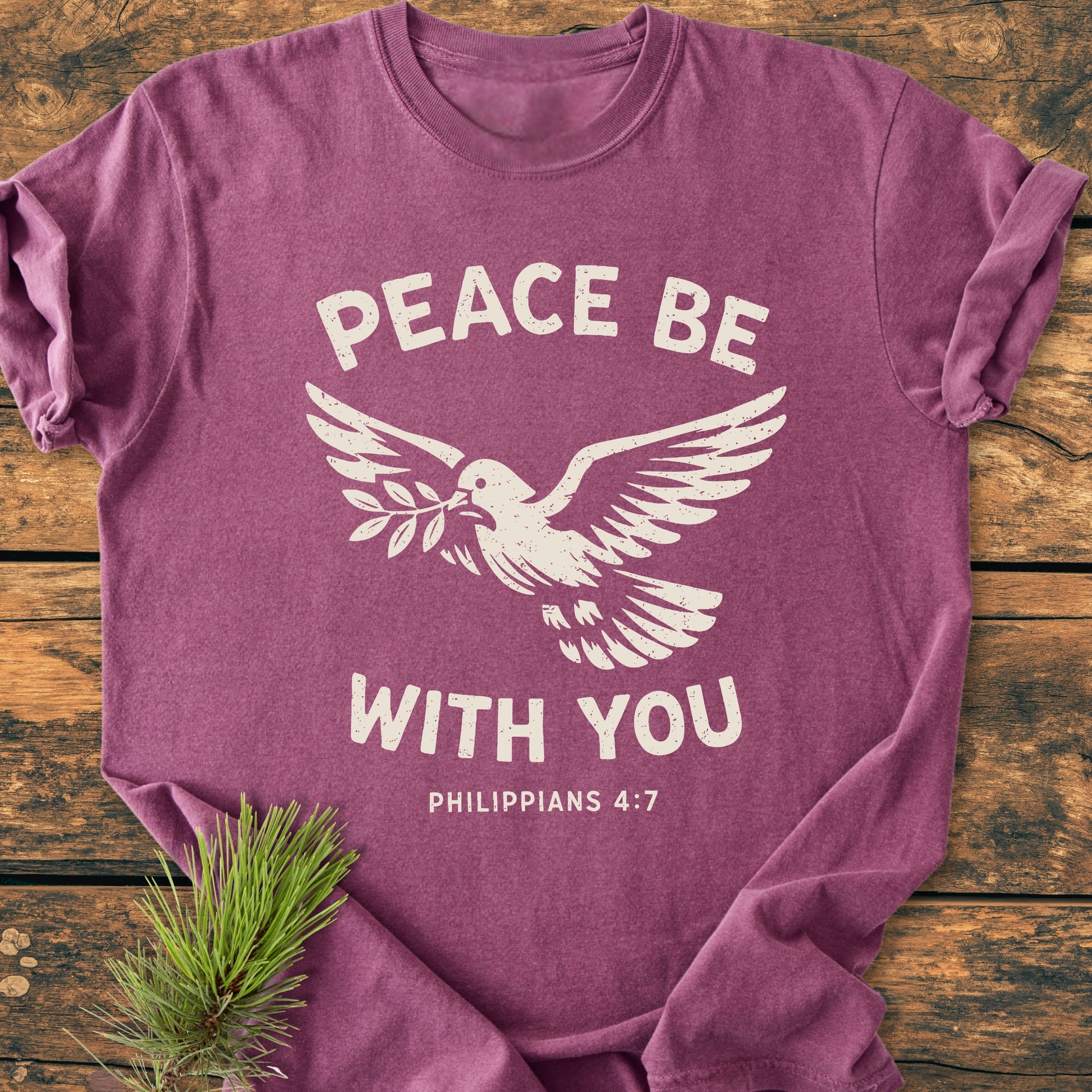 Peace Be With You Philippians 4:7 - Vintage Wash Tee in heathered plum, featuring a white dove and text design. Laid on wood, showcasing its relaxed fit and soft-washed, garment-dyed cotton.