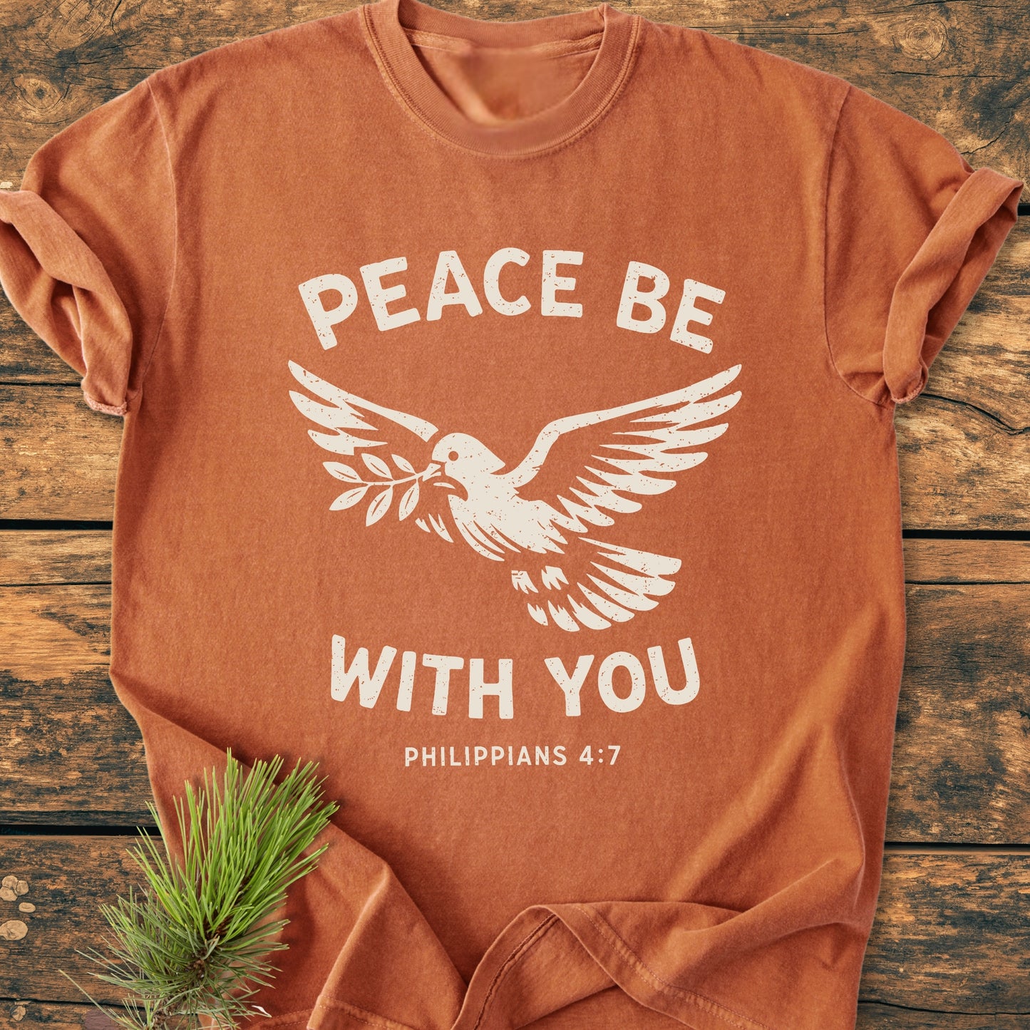 Peace Be With You Philippians 4:7 - Vintage Wash Tee features a burnt-orange crew-neck, dove design with text, laid on wood, highlighting its soft, durable, and relaxed garment-dyed fabric.