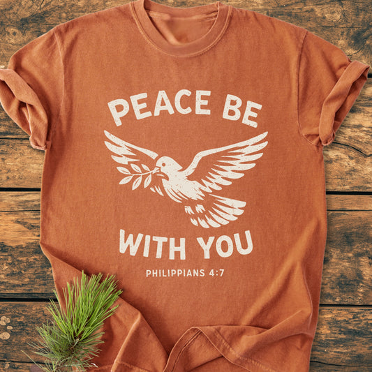 Peace Be With You Philippians 4:7 - Vintage Wash Tee features a burnt-orange crew-neck, dove design with text, laid on wood, highlighting its soft, durable, and relaxed garment-dyed fabric.