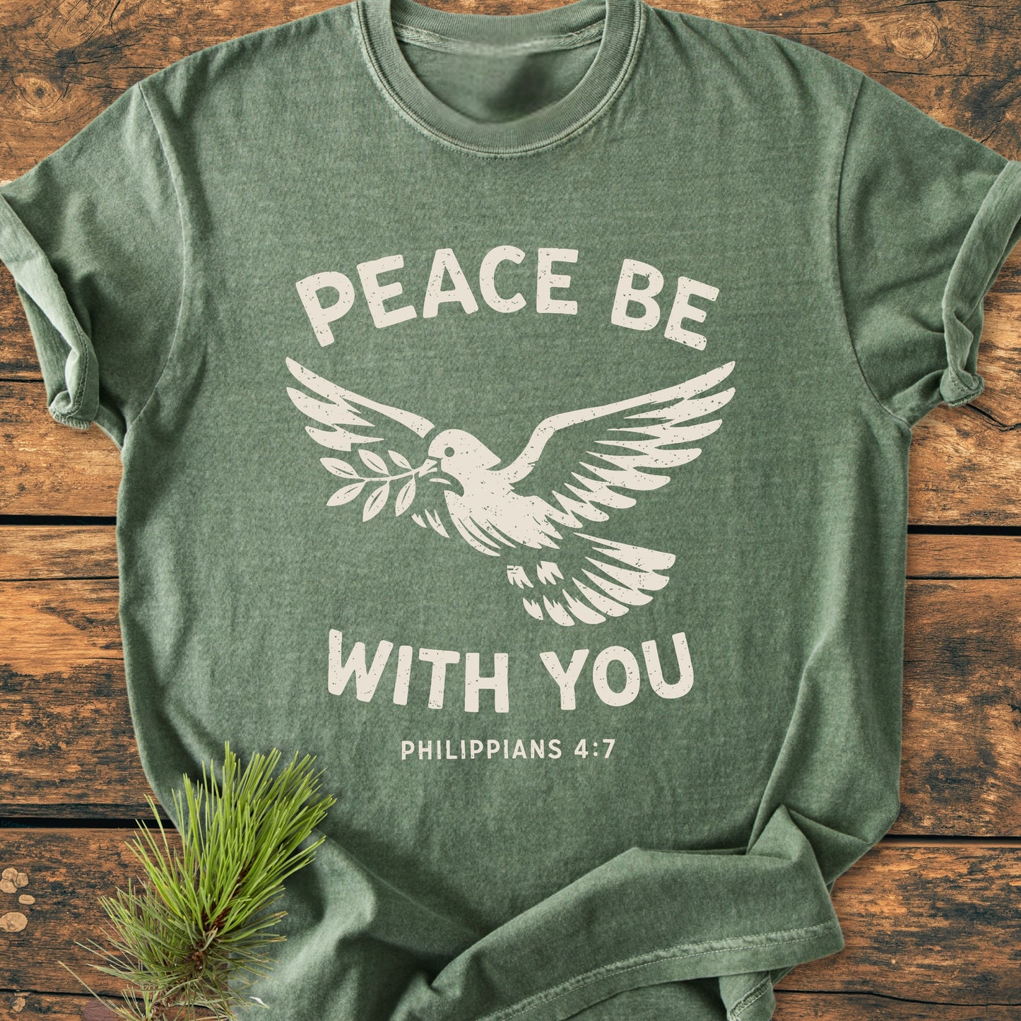 Peace Be With You Philippians 4:7 - Vintage Wash Tee; features a stylized dove and text on a heather-green shirt, displayed on a rustic wooden surface with a pine sprig accent.