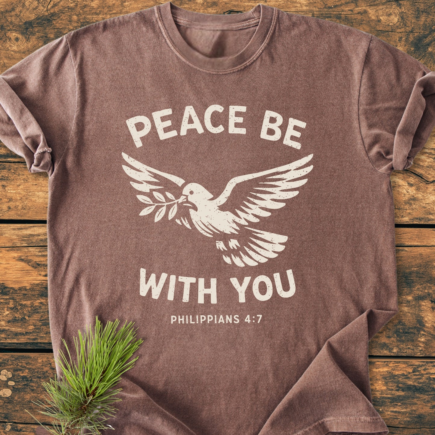 Brown Peace Be With You Philippians 4:7 T-shirt with a stylized dove and olive branch design, laid on wood, featuring soft-washed, garment-dyed fabric, relaxed fit, and durable stitching.
