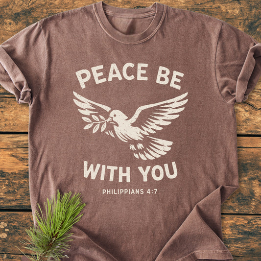 Brown Peace Be With You Philippians 4:7 T-shirt with a stylized dove and olive branch design, laid on wood, featuring soft-washed, garment-dyed fabric, relaxed fit, and durable stitching.