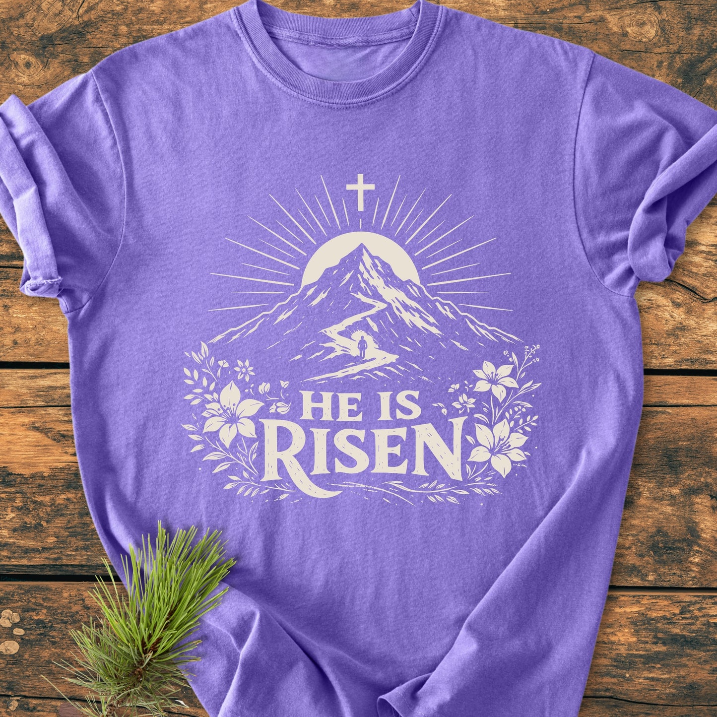 He Is Risen - Vintage Wash Tee features a mountain design with a sun and cross, framed by florals and HE IS RISEN text, laid on wood with an evergreen sprig beside it.
