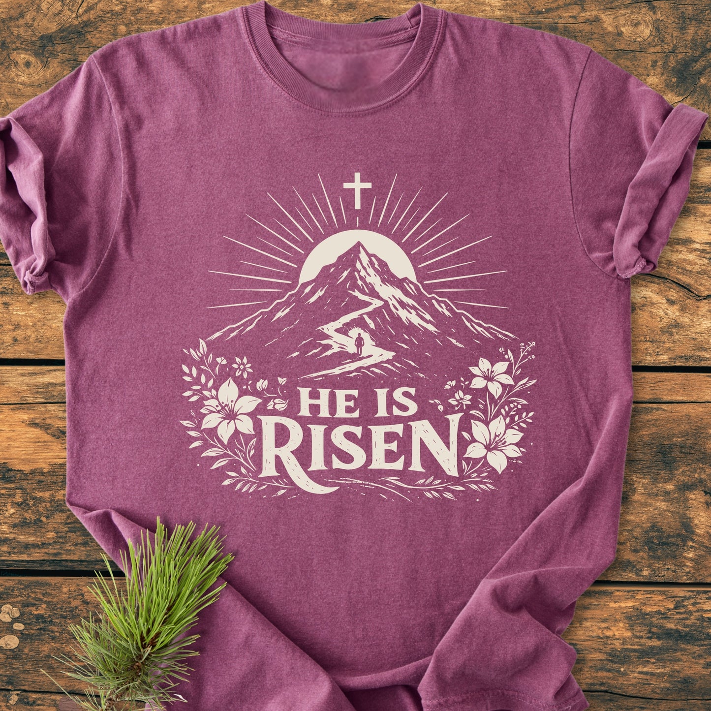 He Is Risen - Vintage Wash Tee: Laid-flat, heathered mauve tee with stylized mountain, sun, and floral design, featuring HE IS RISEN text, displayed on a rustic wooden surface.
