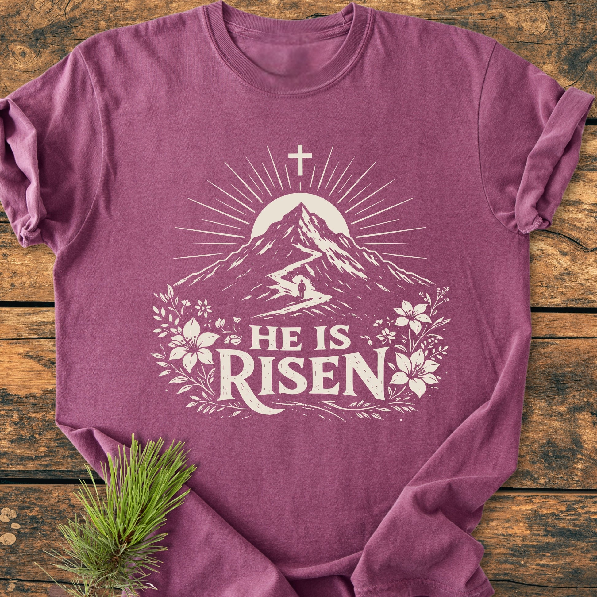He Is Risen - Vintage Wash Tee: Laid-flat, heathered mauve tee with stylized mountain, sun, and floral design, featuring HE IS RISEN text, displayed on a rustic wooden surface.