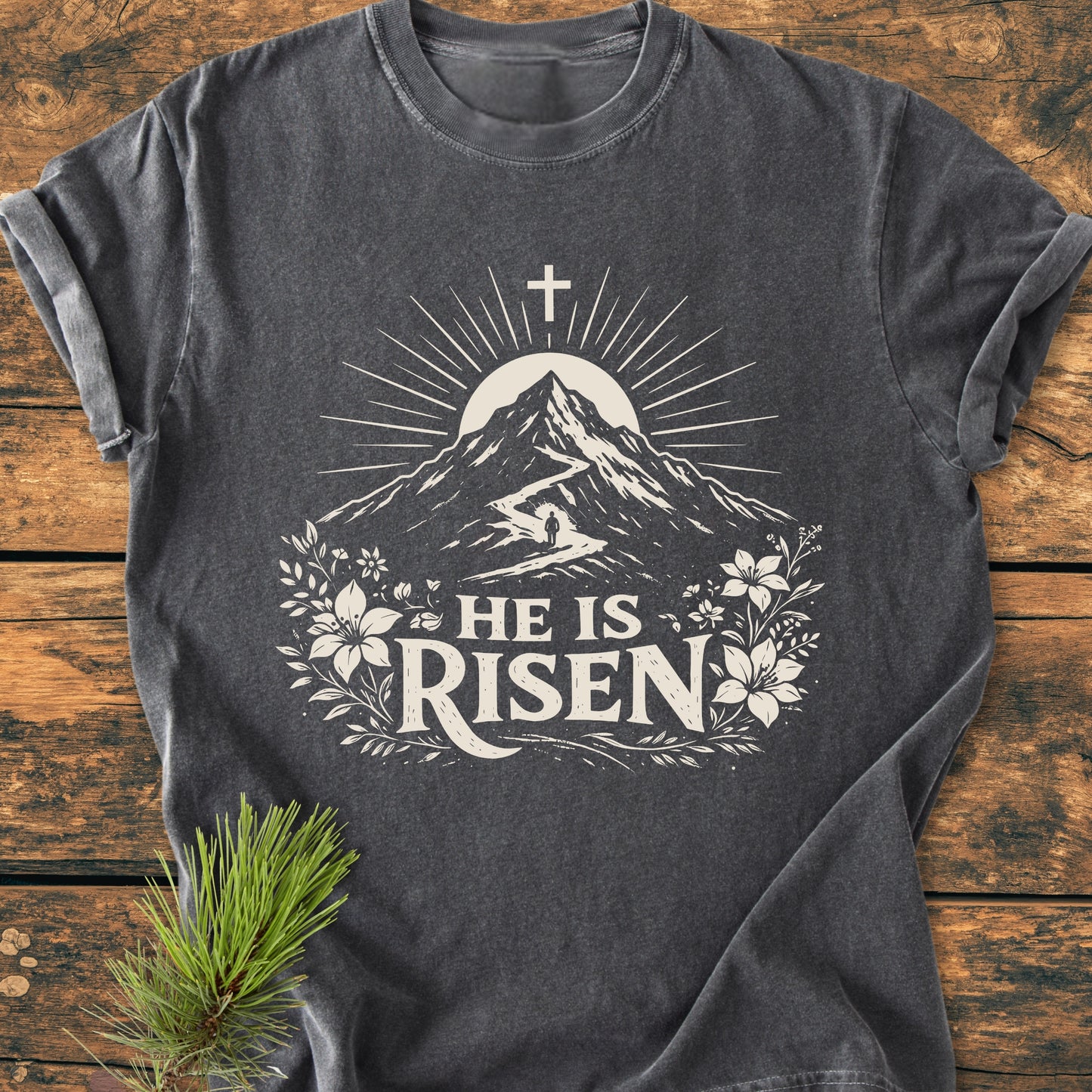 He Is Risen - Vintage Wash Tee featuring a stylized mountain and cross graphic with HE IS RISEN text, laid on wood with a pine twig accent, highlighting the T-shirt's comfort and design.
