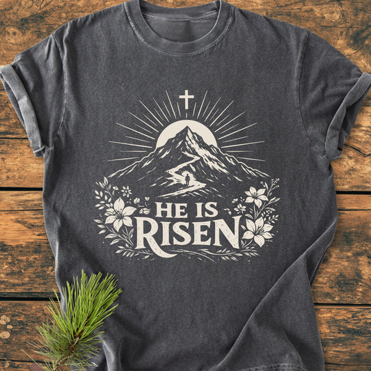 He Is Risen - Vintage Wash Tee featuring a stylized mountain and cross graphic with HE IS RISEN text, laid on wood with a pine twig accent, highlighting the T-shirt's comfort and design.