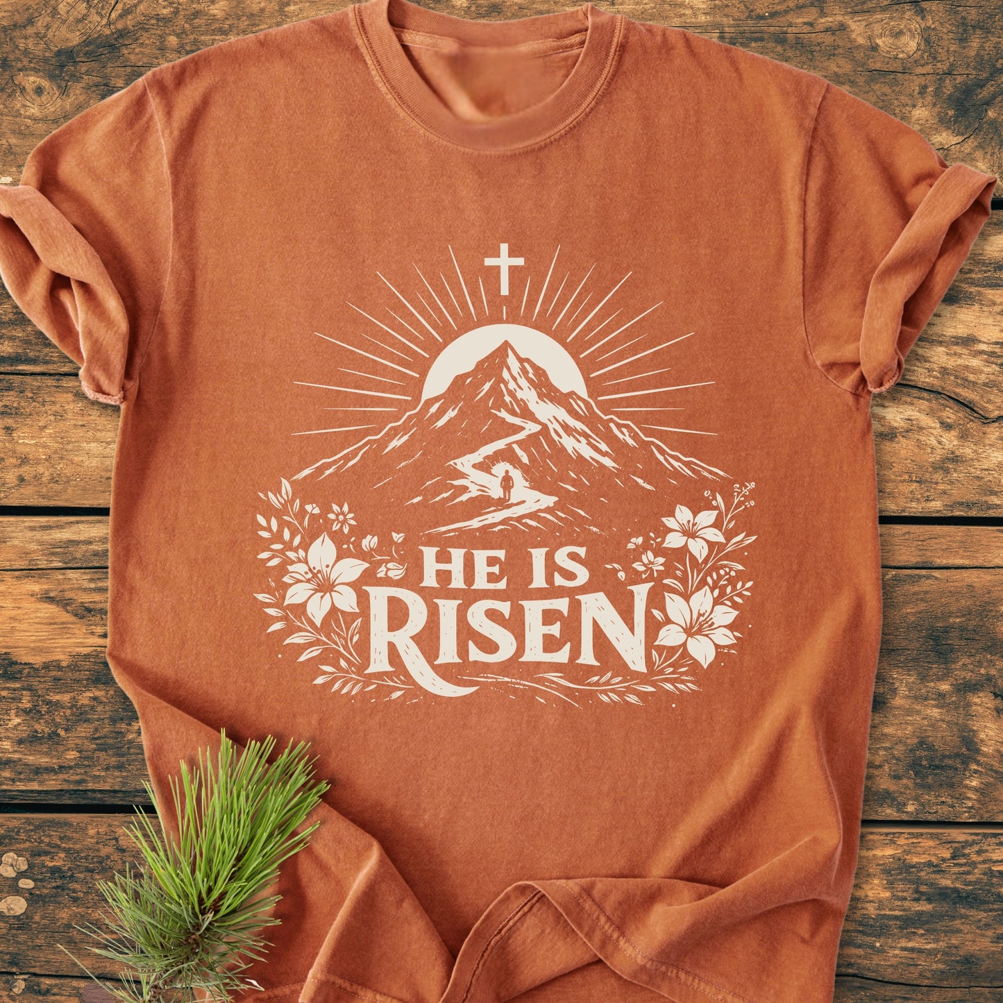 He Is Risen - Vintage Wash Tee: Burnt-orange T-shirt with a white screen-print of a mountain, sun, hiker, and floral design, featuring the text HE IS RISEN on a rustic wooden backdrop.