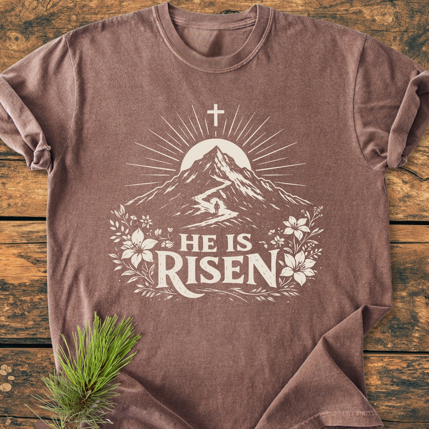 He Is Risen - Vintage Wash Tee: Brown T-shirt featuring a stylized Christian design with a sun, mountain path, figure, cross, and lilies, accompanied by HE IS RISEN text, laid flat on wood.