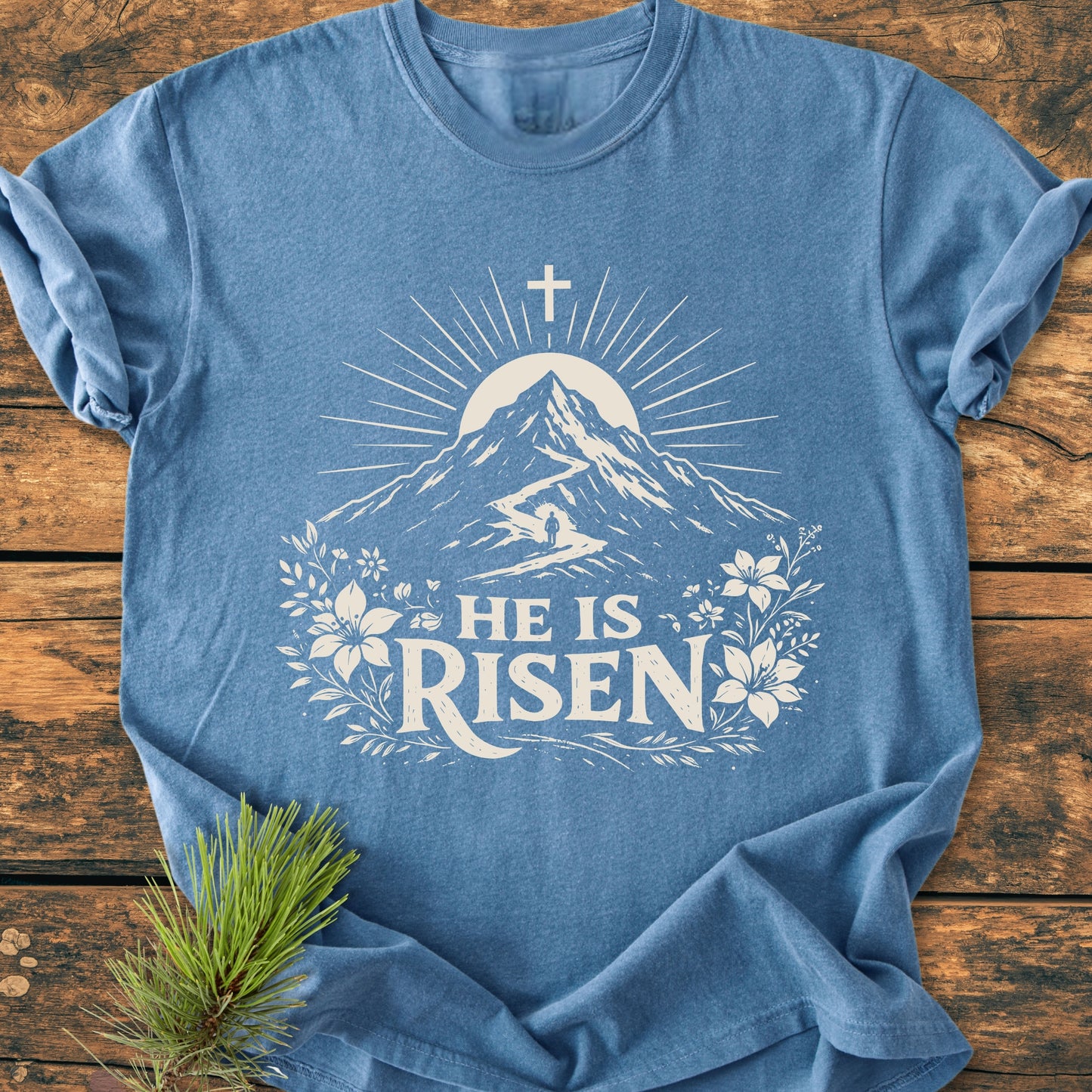 He Is Risen - Vintage Wash Tee features a mountain graphic with sunbeams, climber, and cross, framed by florals on a soft, relaxed-fit, heather-blue Comfort Colors T-shirt.