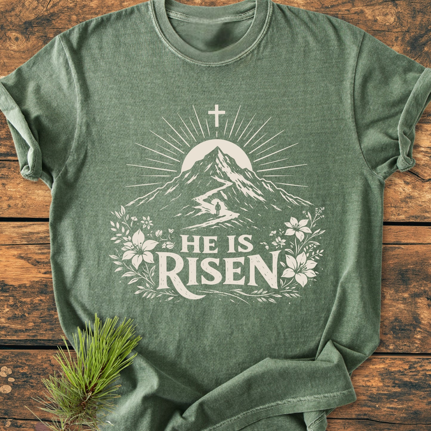 He Is Risen - Vintage Wash Tee laid on wood, featuring a sunrise, mountain, path with climber, and flowers. 'HE IS RISEN' text beneath. Pine sprig accents this heather-green, cozy, and durable shirt.