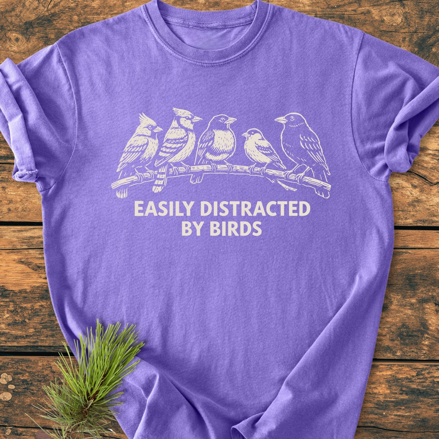 Easily Distracted By Birds - Vintage Wash Tee featuring white birds on a branch and slogan, laid flat on wood with pine sprig, showcasing its soft, relaxed fit and durable stitching.
