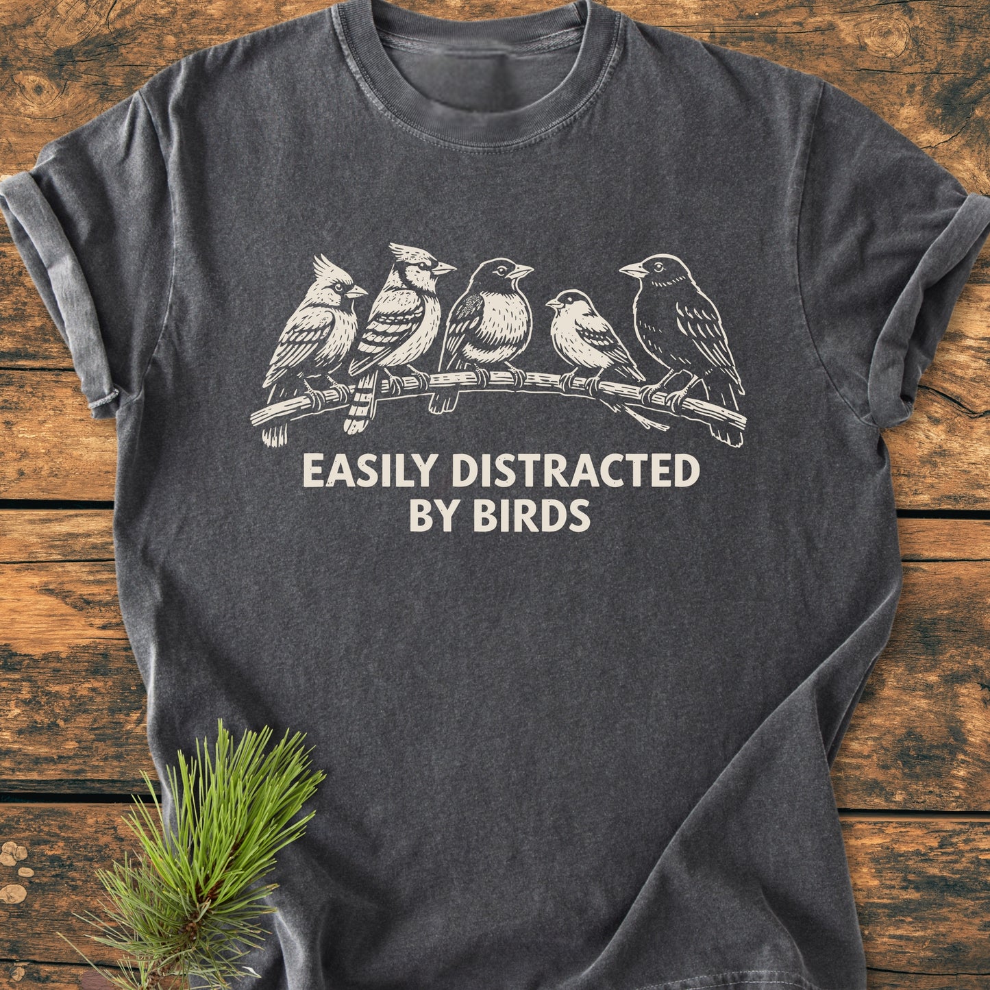 Easily Distracted By Birds - Vintage Wash Tee: Charcoal-gray T-shirt with five stylized birds on a branch and slogan. Laid flat on wood, showcasing soft, relaxed fit and durable stitching.