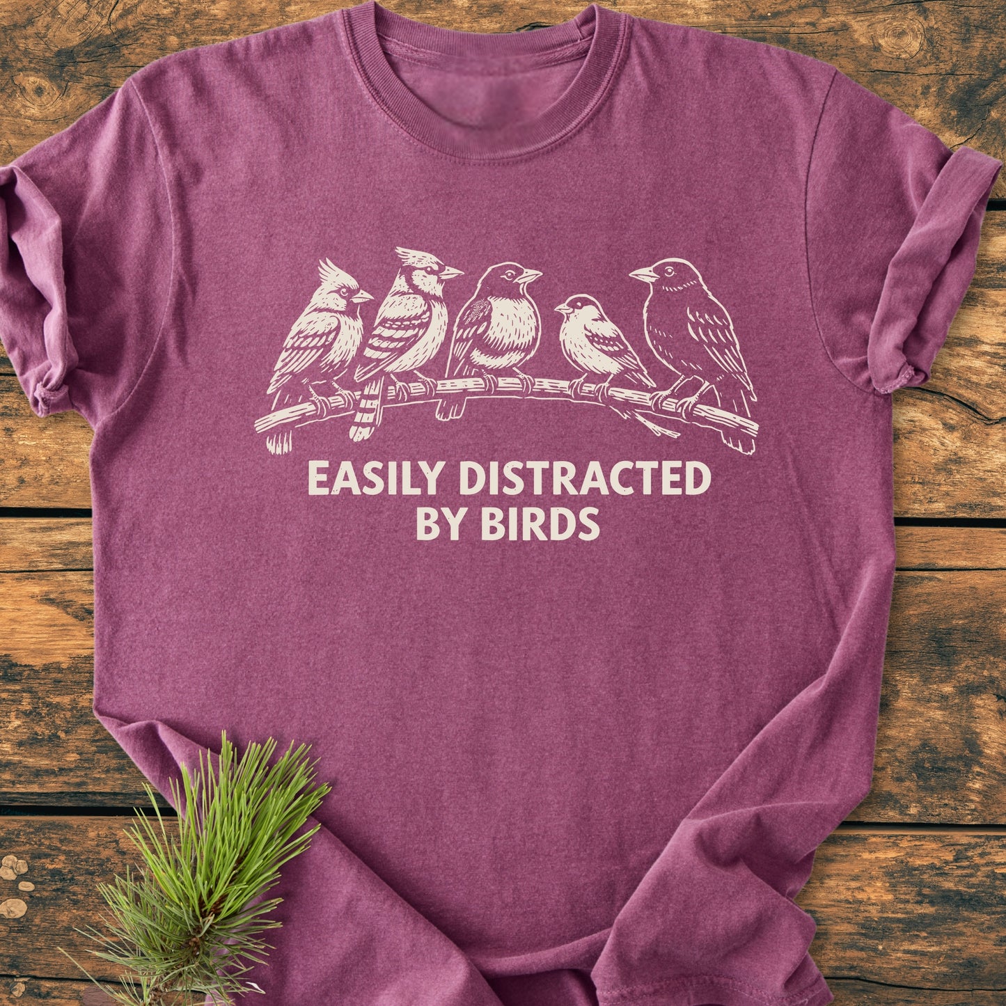 Easily Distracted By Birds - Vintage Wash Tee features a heather-purple color with five birds on a branch and text. Laid flat on rustic wood, highlighting its relaxed fit and soft-washed fabric.