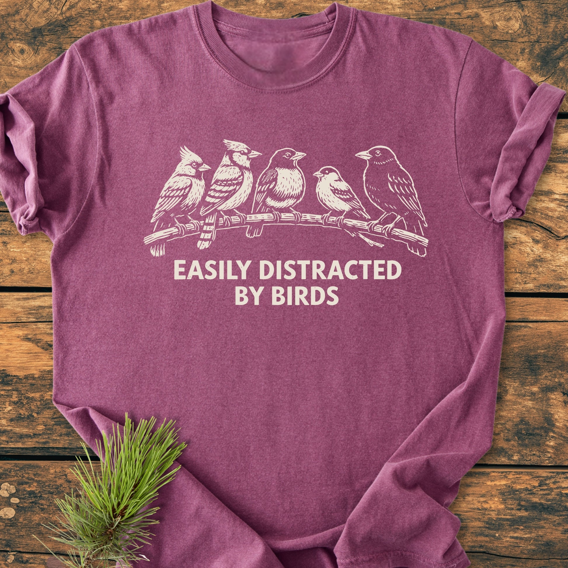 Easily Distracted By Birds - Vintage Wash Tee features a heather-purple color with five birds on a branch and text. Laid flat on rustic wood, highlighting its relaxed fit and soft-washed fabric.
