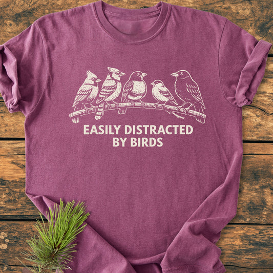 Easily Distracted By Birds - Vintage Wash Tee features a heather-purple color with five birds on a branch and text. Laid flat on rustic wood, highlighting its relaxed fit and soft-washed fabric.