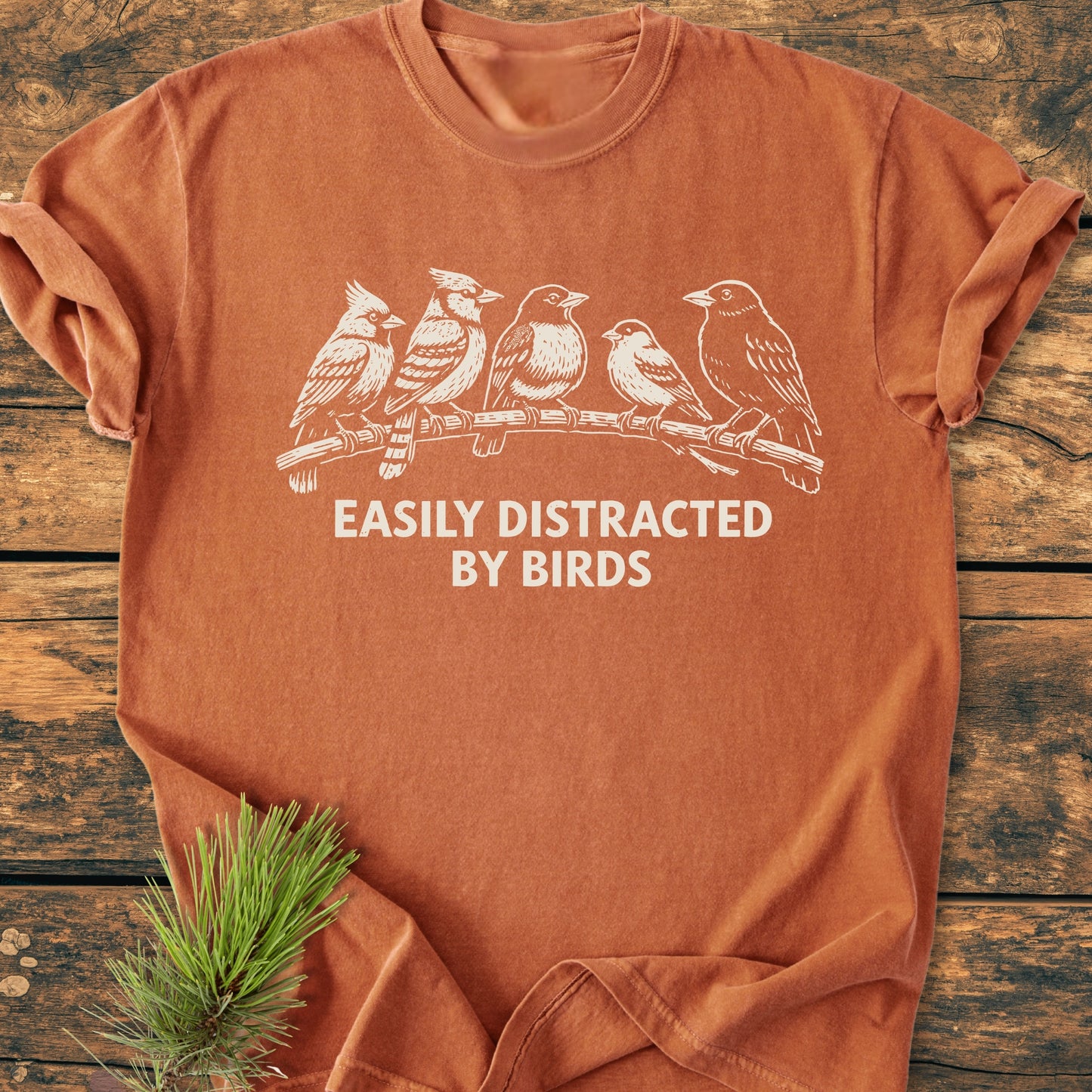 Easily Distracted By Birds - Vintage Wash Tee: Rust-orange, short-sleeve T-shirt with five birds design and slogan, laid flat on wood, highlighting its relaxed, cozy fit and durable, garment-dyed fabric.
