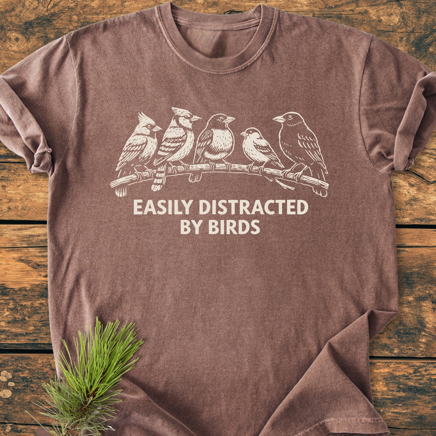 Easily Distracted By Birds - Vintage Wash Tee features a white bird line-drawing and slogan on a soft, brown heathered T-shirt, displayed flat-lay with a pine sprig accentuating its rustic appeal.