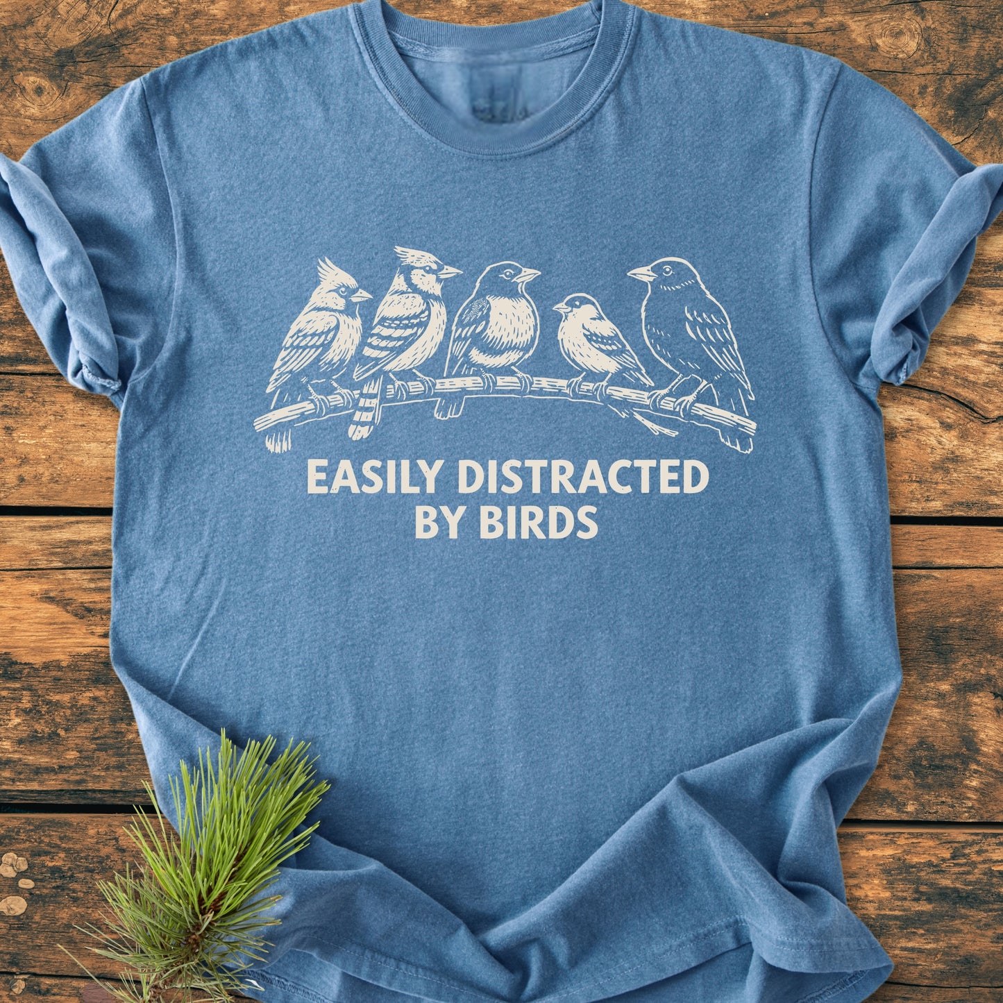 Easily Distracted By Birds - Vintage Wash Tee featuring a white songbird illustration and slogan on a blue heather-style background, laid on wood with a pine sprig accent.