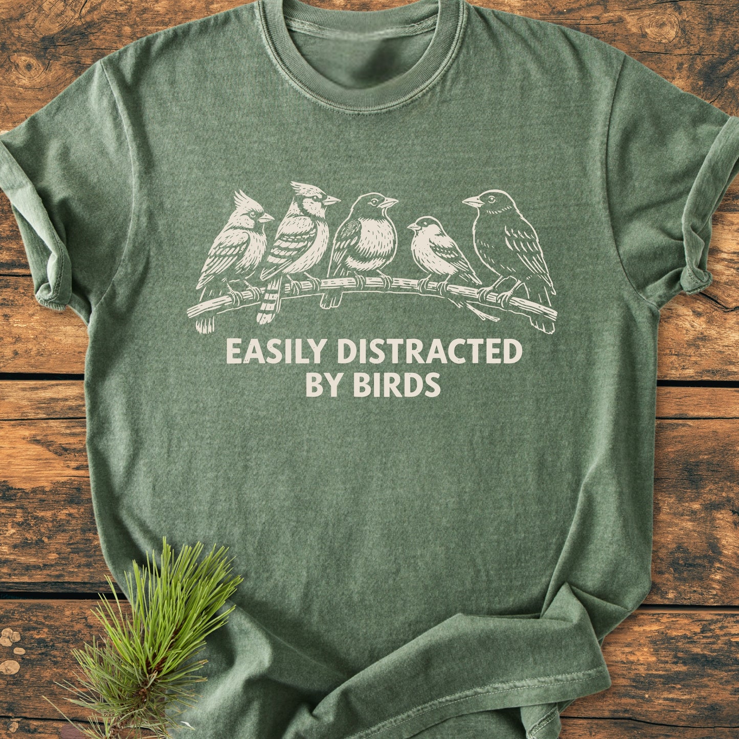 Heather-green Easily Distracted By Birds - Vintage Wash Tee featuring a white line-drawing of songbirds and slogan, displayed on wood with a pine sprig, showcasing its relaxed fit and soft texture.