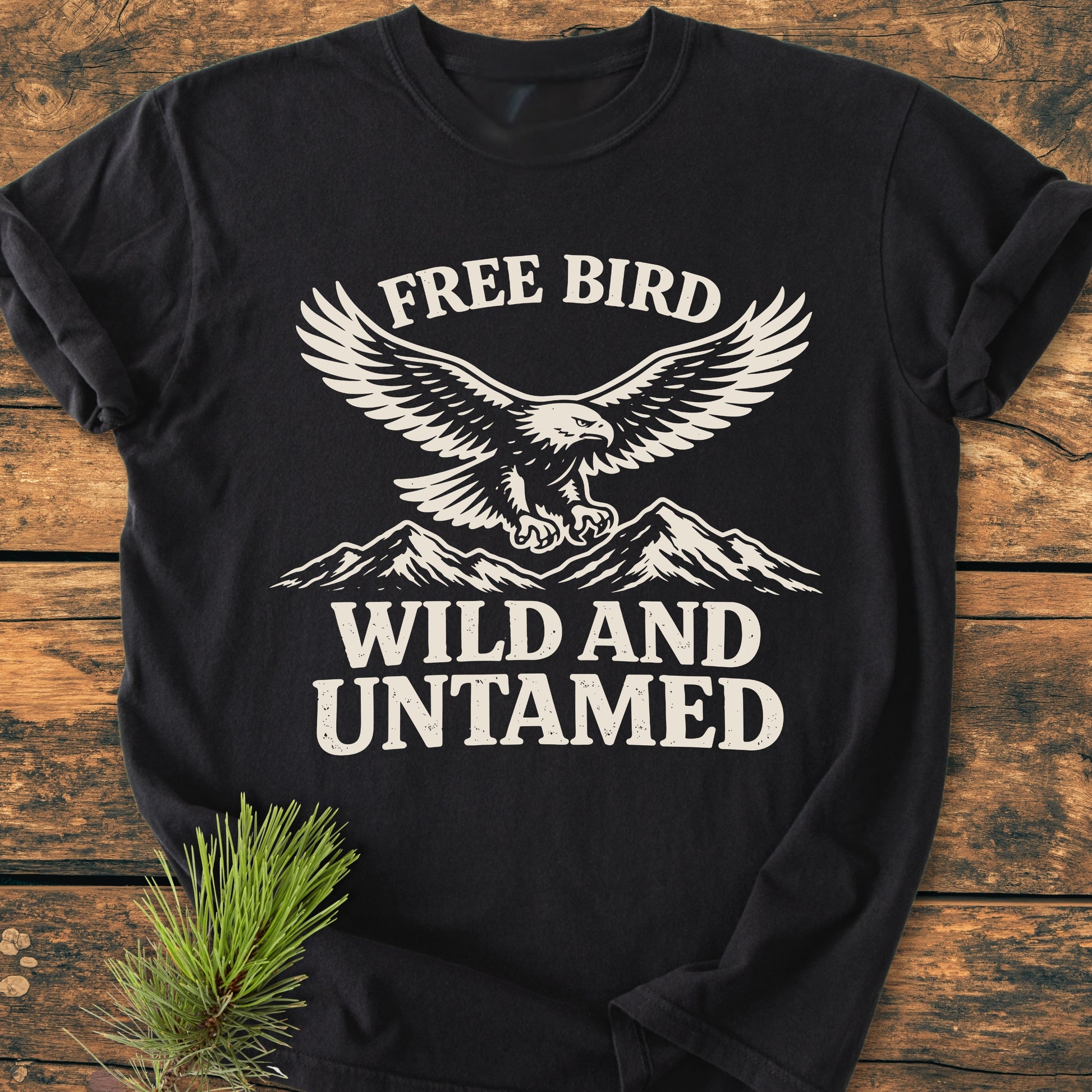 Free Bird - Vintage Wash Tee features a white graphic of a soaring eagle over mountains with “FREE BIRD” and “WILD AND UNTAMED” text, laid on a wooden surface with a pine sprig.