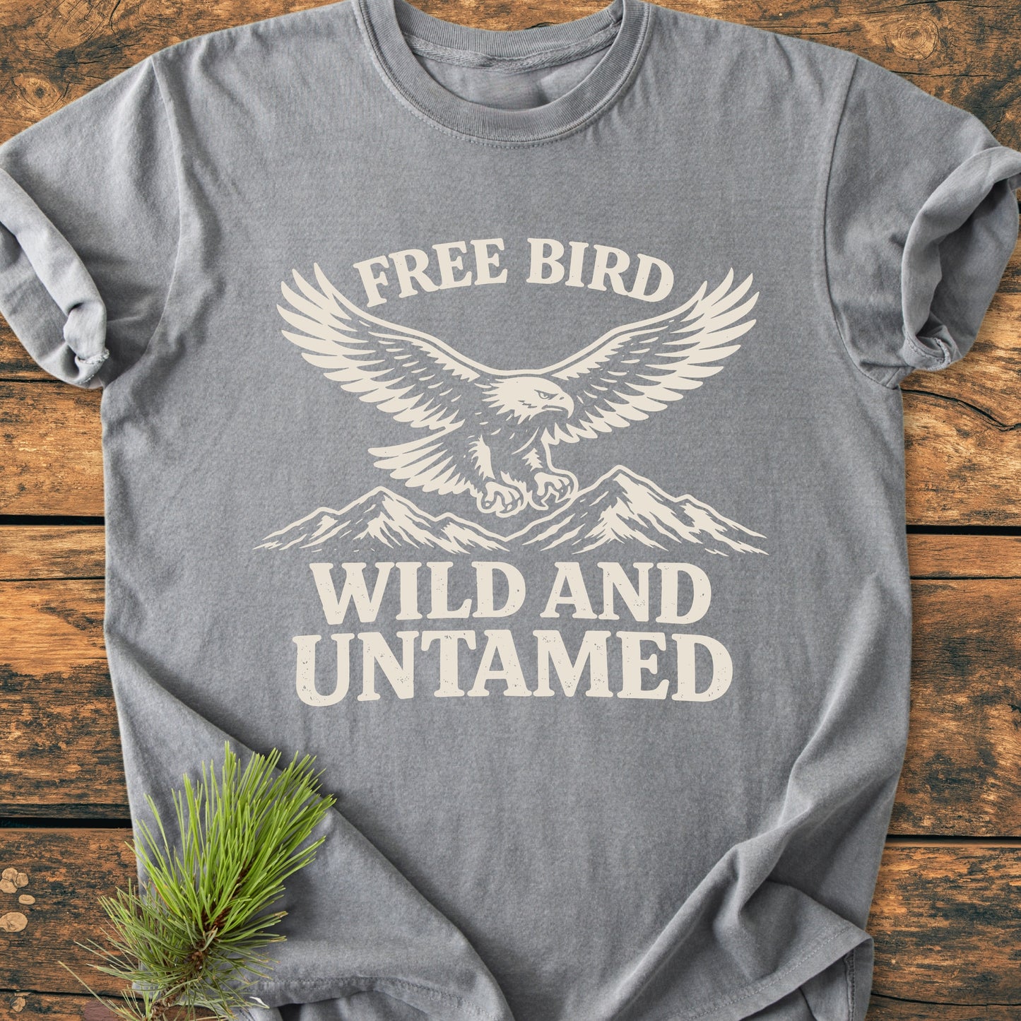 Free Bird - Vintage Wash Tee features a heather-gray, short-sleeve design with an eagle graphic and the phrases “FREE BIRD” and “WILD AND UNTAMED,” laid flat on a wooden surface.