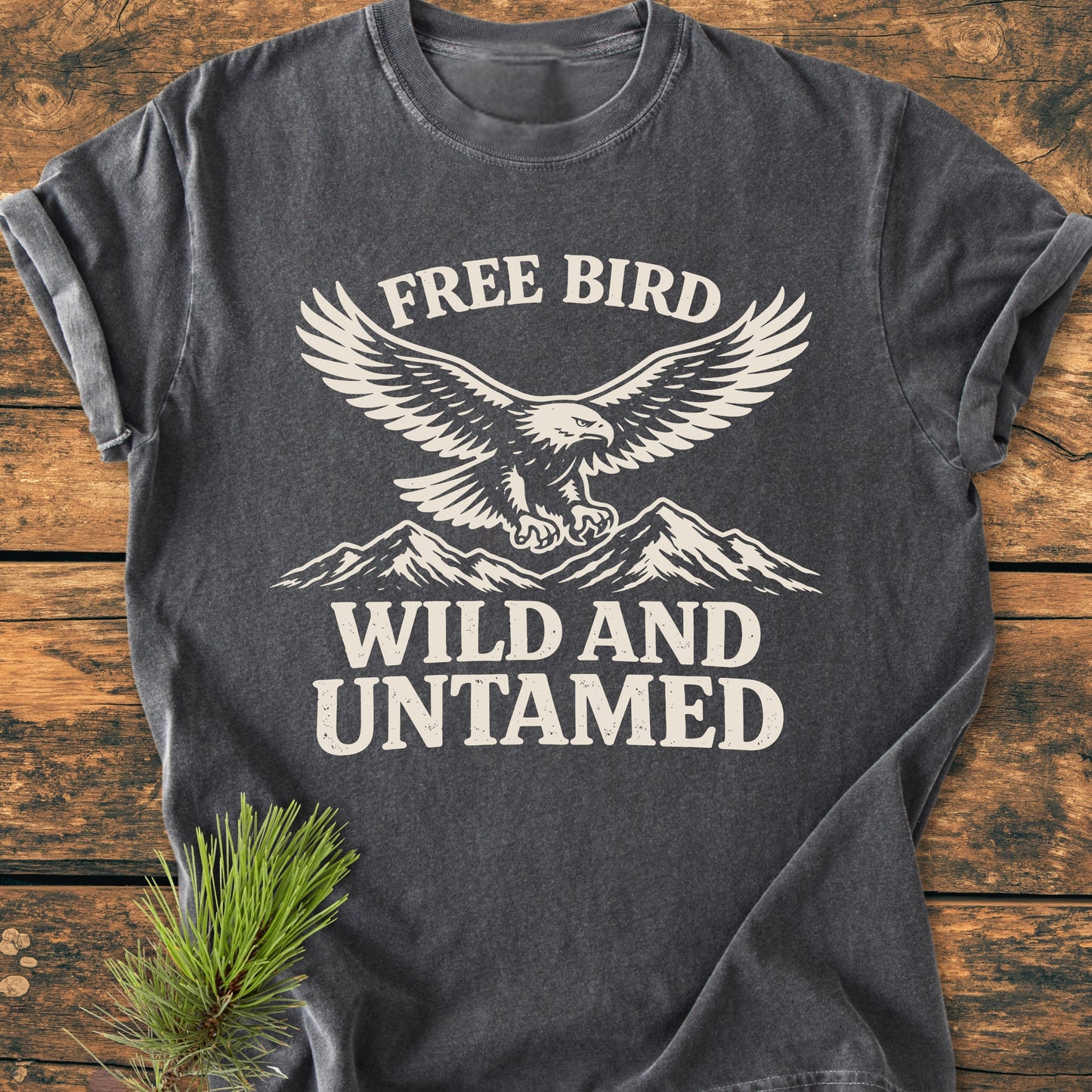 Free Bird - Vintage Wash Tee featuring a stylized eagle over mountains with “FREE BIRD” and “WILD AND UNTAMED” text, displayed on a wooden surface with a pine sprig beside it.