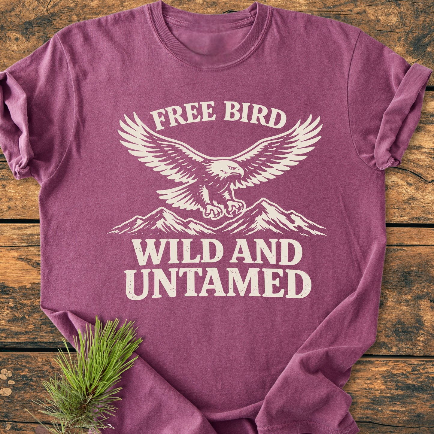 Free Bird - Vintage Wash Tee: Heathered purple T-shirt featuring a cream eagle graphic with FREE BIRD and WILD AND UNTAMED text, displayed on a wooden surface with a pine sprig accent.