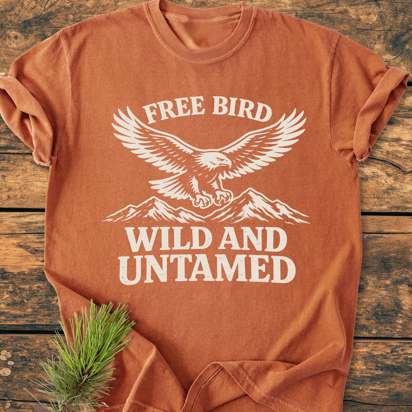 Free Bird - Vintage Wash Tee featuring a stylized eagle and mountains print, laid flat on a wooden surface with a pine sprig beside it. Cozy, durable, and relaxed fit.