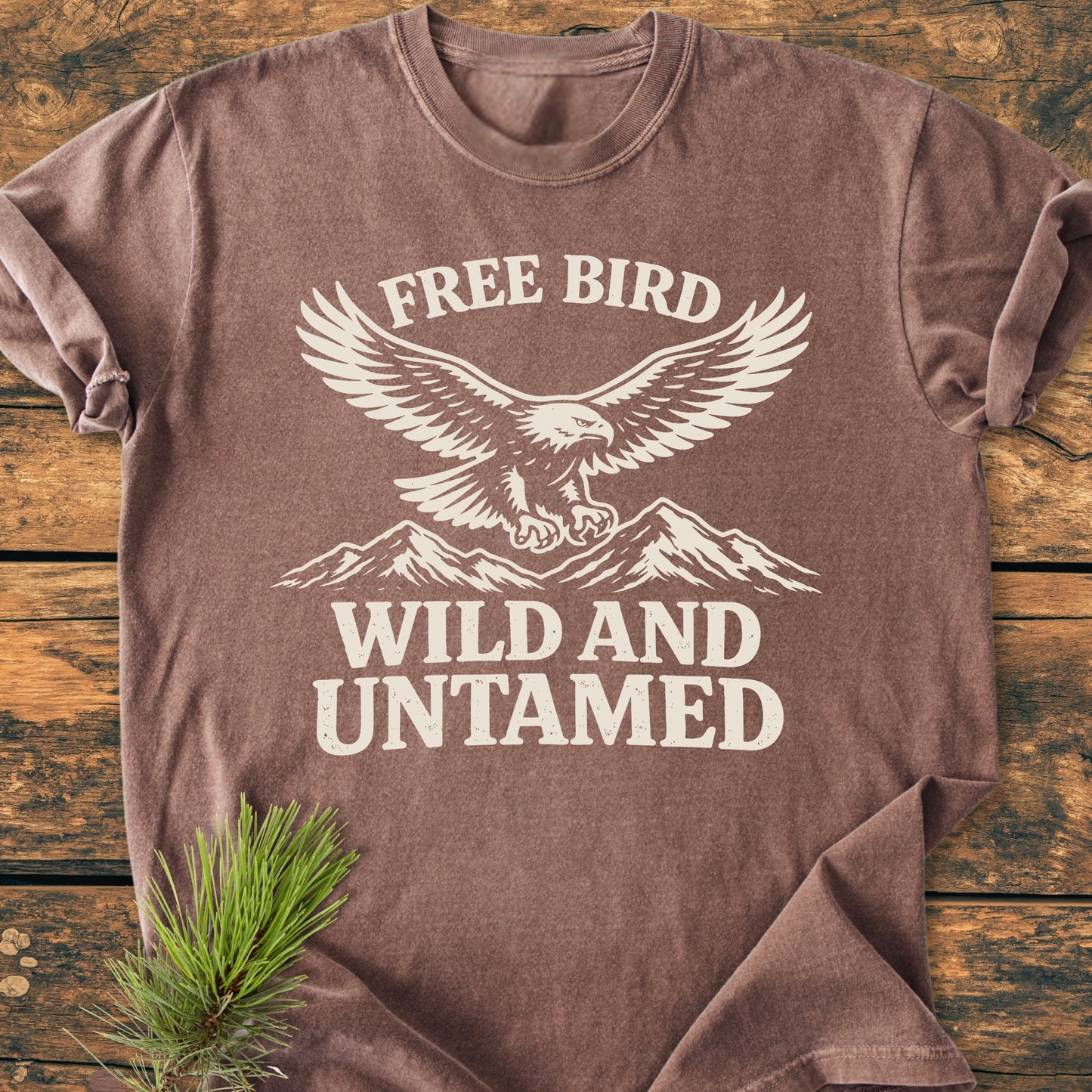 Free Bird - Vintage Wash Tee featuring an off-white eagle and mountain design with slogans, laid on a wooden surface, highlighting its relaxed fit and soft, durable 100% ring-spun cotton fabric.