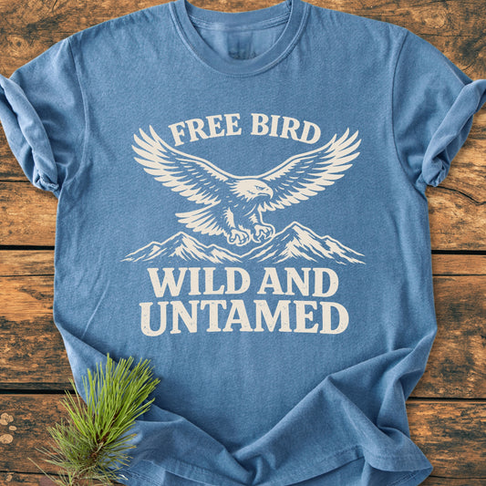 Free Bird - Vintage Wash Tee features a stylized eagle above mountains in off-white print on a heather-blue T-shirt, laid on a rustic surface with a pine needle accent.