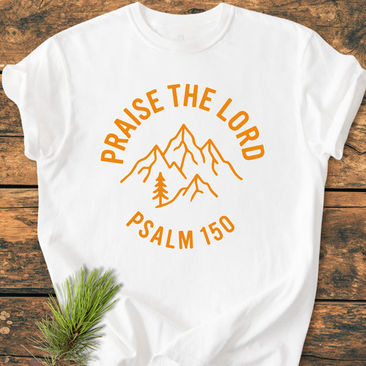 Praise The Lord, Psalm 150 - Vintage Wash Tee: White t-shirt with orange line-drawing of mountains and pine tree, featuring arched text “PRAISE THE LORD” and “PSALM 150” below, laid on wood-grain surface.