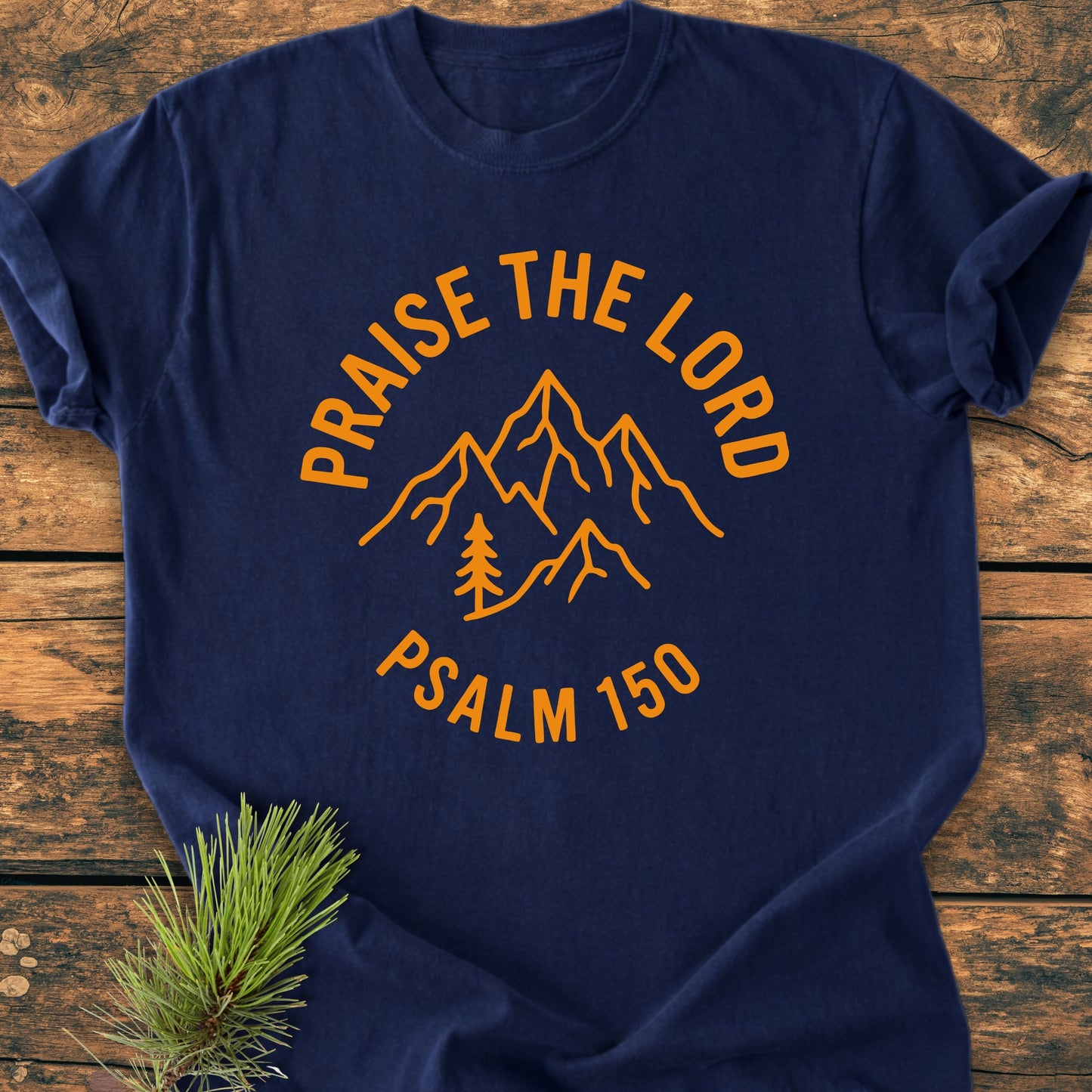 Praise The Lord, Psalm 150 - Vintage Wash Tees features a navy-blue shirt with bold orange mountain and tree design, highlighting PRAISE THE LORD and PSALM 150, laid on wooden surface.