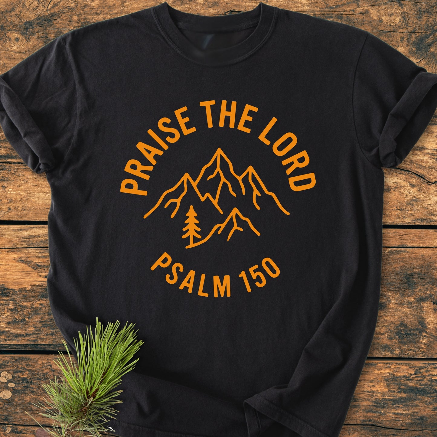 Praise The Lord, Psalm 150 - Vintage Wash Tee: A black T-shirt with a mountain range and pine tree graphic, featuring “PRAISE THE LORD” above and “PSALM 150” below, on a wooden surface.