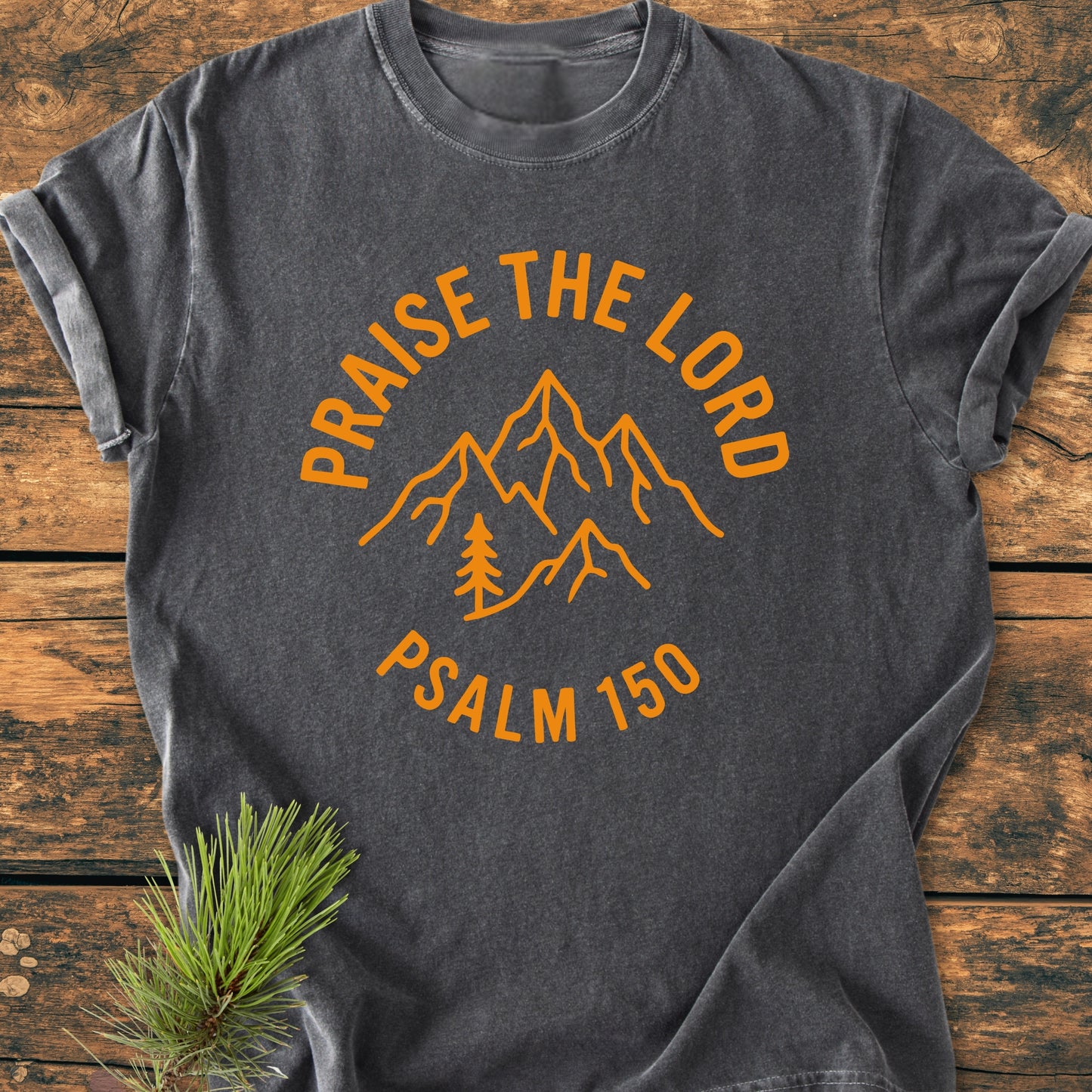 Praise The Lord, Psalm 150 - Vintage Wash Tee: Dark-gray T-shirt with mountain-and-tree design and text, laid on wood surface. Features soft, durable, ring-spun cotton with relaxed fit. Pine sprig accent.