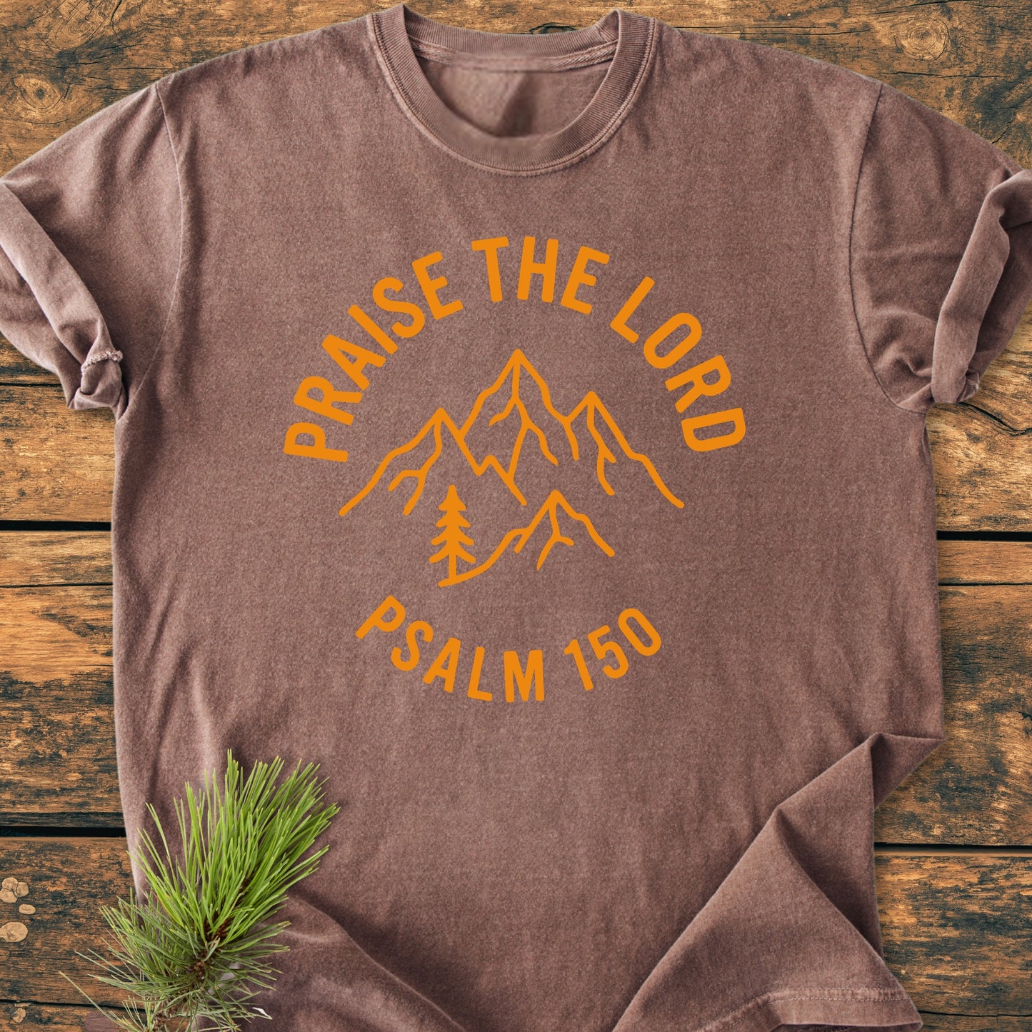 Heathered brown T-shirt featuring Praise The Lord, Psalm 150 with a mountain sketch and pine tree in pumpkin-orange ink, laid flat with a pine branch accent, emphasizing its relaxed, durable design.