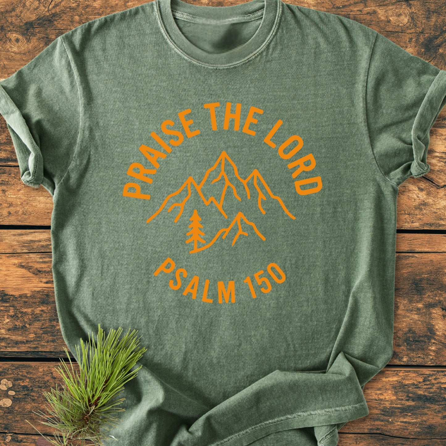 Heather-green T-shirt with Praise The Lord, Psalm 150 design featuring mountain peaks and a pine tree. Styled with pine needles and cones on a wooden surface.