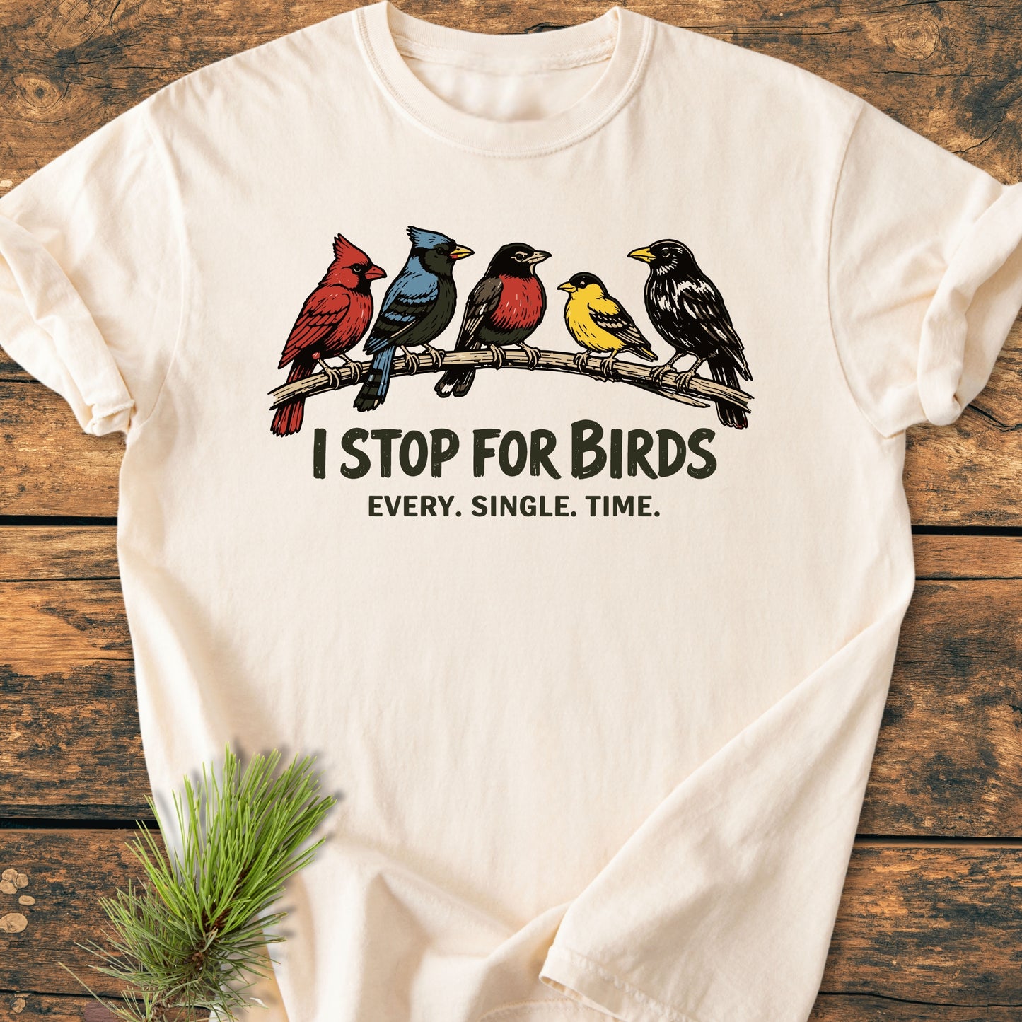 I Stop For Birds - Vintage Wash Tee features five illustrated birds on a branch with bold slogans, laid on a wooden surface, showcasing its relaxed fit and off-white, garment-dyed fabric.